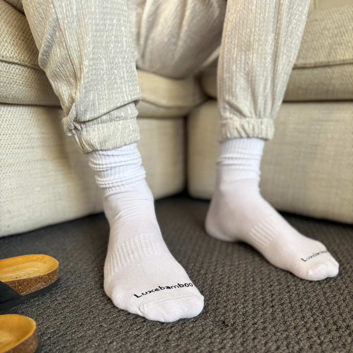 Person wearing white bamboo socks with 'Luxebamboo' branding, sitting on a beige couch.