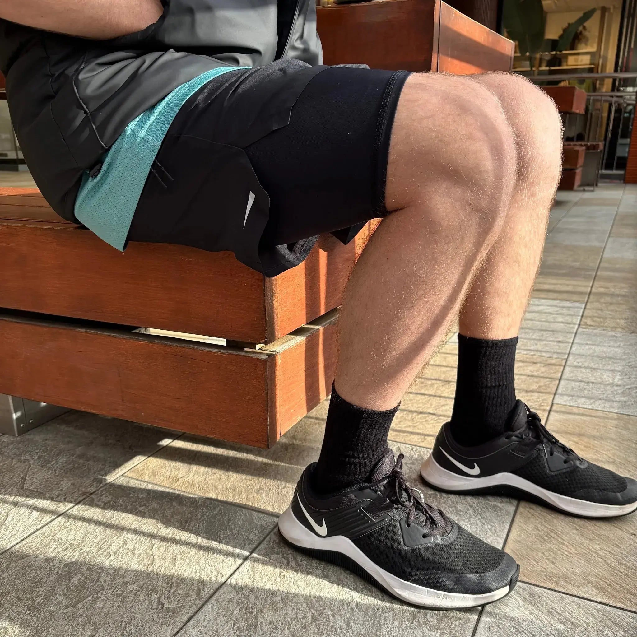 Person wearing black athletic shoes with black bamboo socks and sports shorts sitting on a wooden bench.